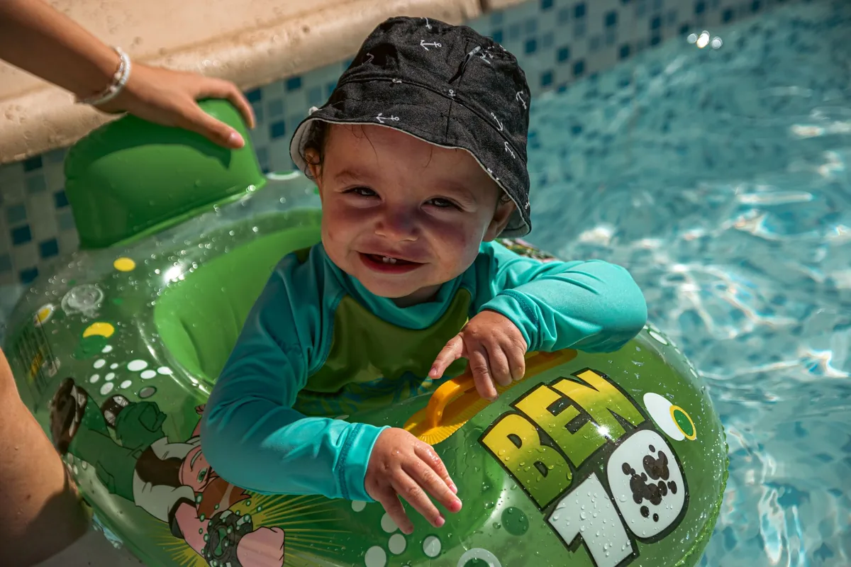 Adorable toddler in sun hat and green swim ring smiles while enjoying a sunny pool day